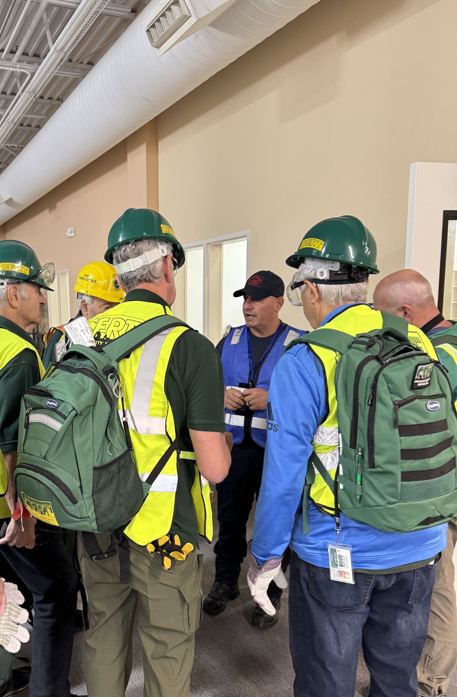 A team in green hard hats and high-visibility vests standing in a briefing huddle, seen from behind, carrying response packs and work gloves