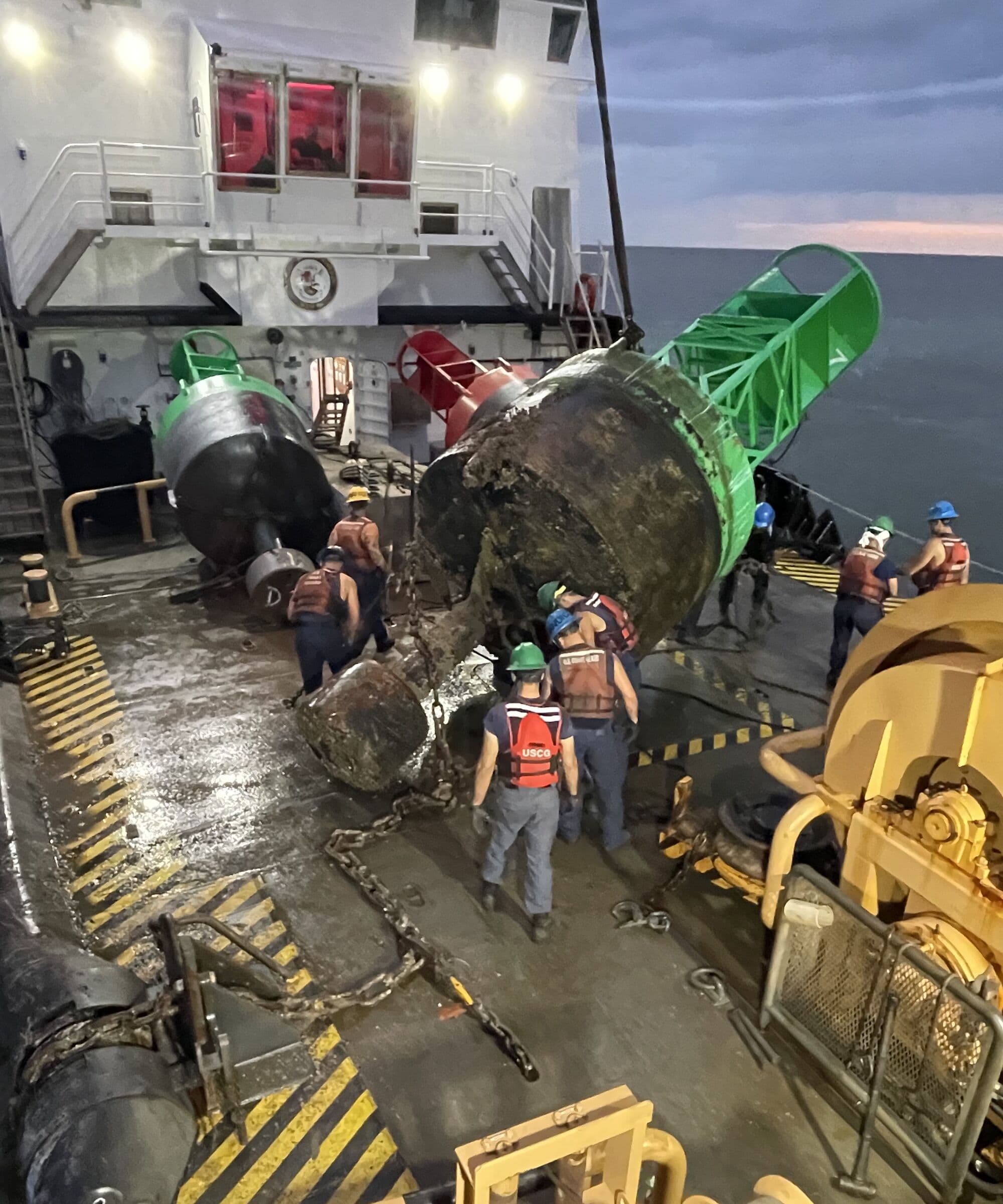 A Coast Guard buoy deck at dusk, crew in hard hats and float coats working a barnacle-covered navigation buoy hanging on a crane wire beside its bright replacement