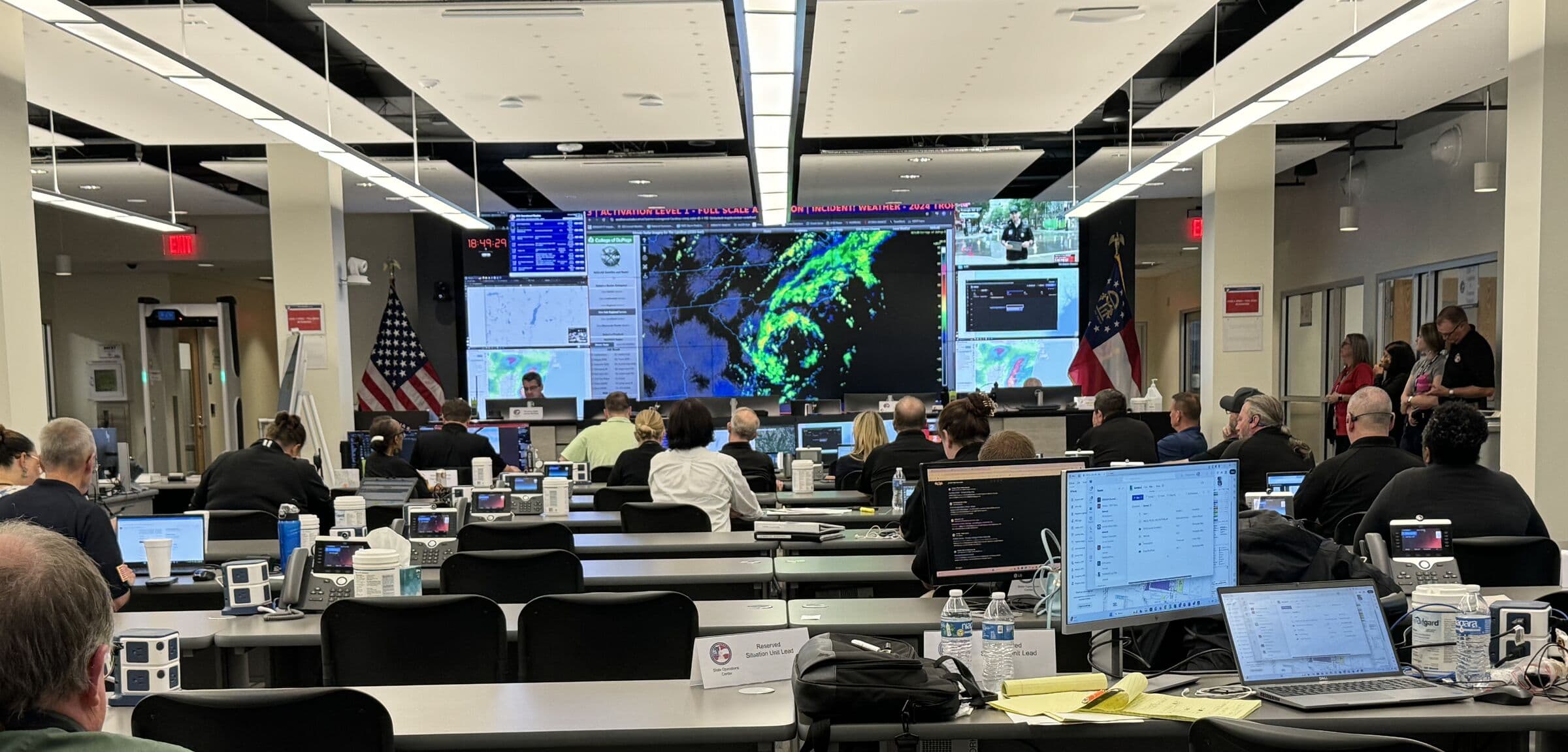 A state emergency operations centre at full activation, staff at rows of desks facing a wall-sized weather radar display