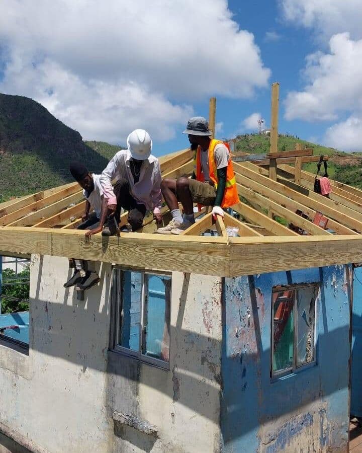 Three workers building the fresh timber roof frame of a small blue and grey house, one in a white hard hat and one in an orange high-visibility vest, with a green hillside behind them